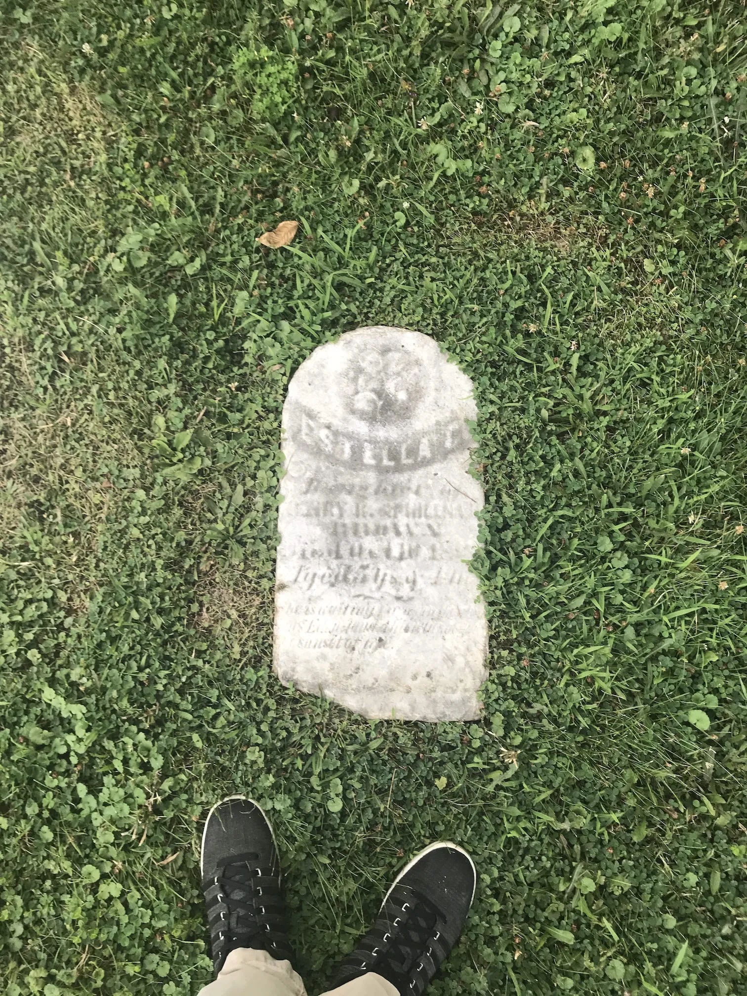 A photo of a really old headstone embedded in the ground at a cemetary in the summer